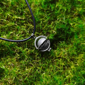 Pode incluir: Um colar de prata com um pingente de globo giratório. O colar é feito de cordão preto e o globo está suspenso em uma moldura em forma de gaiola.