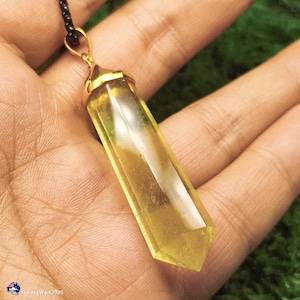 Op de afbeelding: Een gele kristallen hanger met een goudkleurige dop en een zwarte ketting. De kristal is lichtgeel van kleur en is gesneden in een lange, rechthoekige vorm met een puntig uiteinde. De hanger wordt in een hand gehouden.