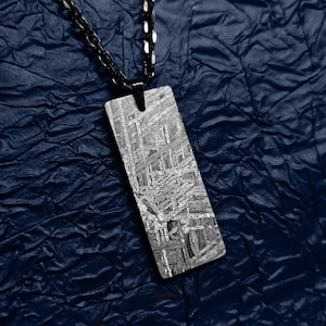 Puede incluir: Un collar con un colgante rectangular de meteorito de color plateado. El colgante tiene una cadena negra y está sobre un fondo azul texturizado.
