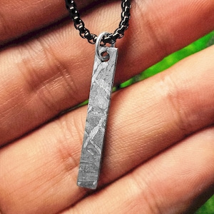 Peut inclure: Un pendentif rectangulaire avec un motif texturé unique est suspendu à une chaîne noire. Le pendentif est de couleur gris clair avec un motif détaillé entrecroisé. La chaîne est de couleur gris foncé. Le bijou est présenté sur un fond flou.