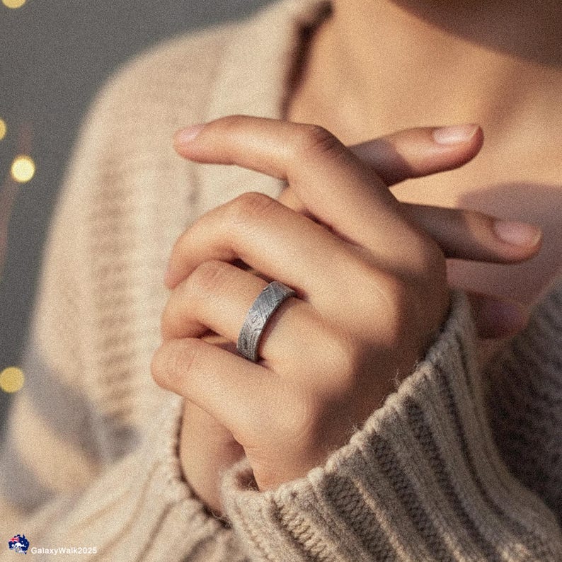 May include: A silver ring with a textured pattern is worn on a person's finger. The ring's design features a series of diagonal lines, creating a unique visual texture. The person is wearing a beige jumper.