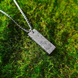 Puede incluir: Un collar de plata con un colgante rectangular hecho de metal de meteorito. El colgante tiene una superficie texturizada única con un patrón de líneas y remolinos. El collar está fotografiado sobre un lecho de musgo verde.