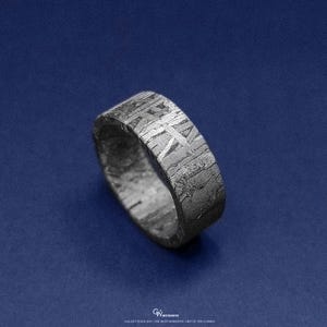 May include: A textured, dark grey ring with a distinctive pattern, set against a dark blue background. The ring's surface has a cross-hatched design. The ring appears to be made of metal. The ring is a band style.