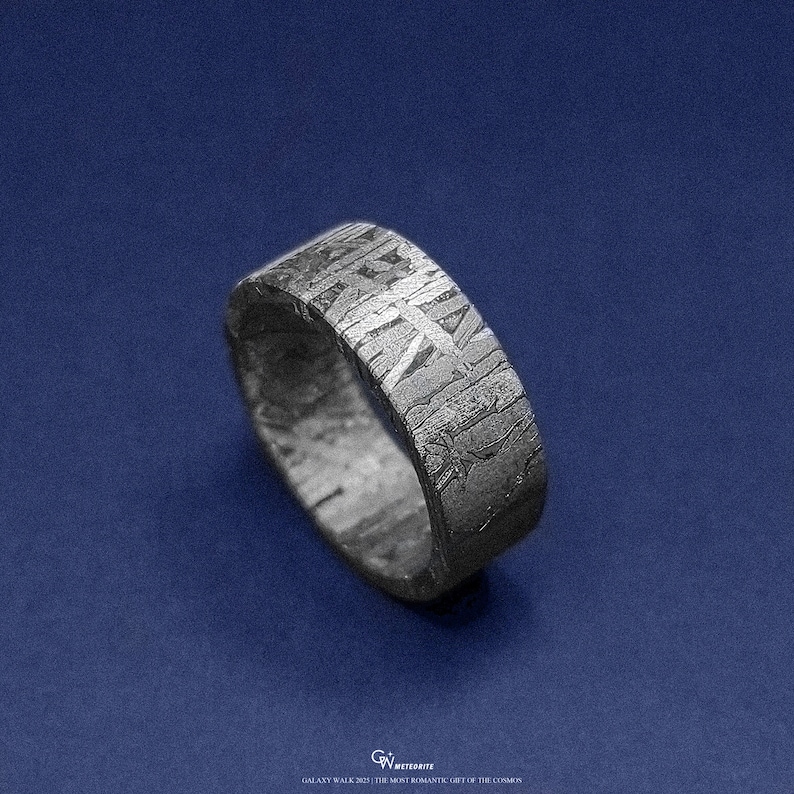 May include: A textured, dark grey ring with a distinctive pattern, set against a dark blue background. The ring's surface has a cross-hatched design. The ring appears to be made of metal. The ring is a band style.