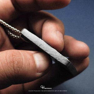May include: A rectangular pendant with a patterned surface, suspended from a silver chain. The pendant is held in a hand against a blue background. Text at the bottom reads "GALAXY WALK 2025 | THE MOST ROMANTIC GIFT OF THE COSMOS."