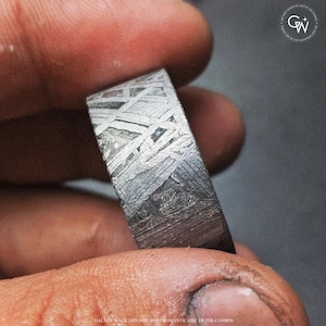 May include: Close-up of a rectangular ring with a distinctive pattern, crafted from a dark, metallic substance. The ring features a unique, textured design with intersecting lines and a polished finish. The ring is held between fingers.