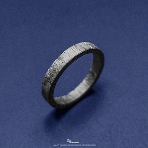 May include: A dark grey ring with a textured surface and a distinctive pattern, set against a deep blue backdrop. The ring's design has a unique, almost metallic appearance. The ring is a piece of jewellery.