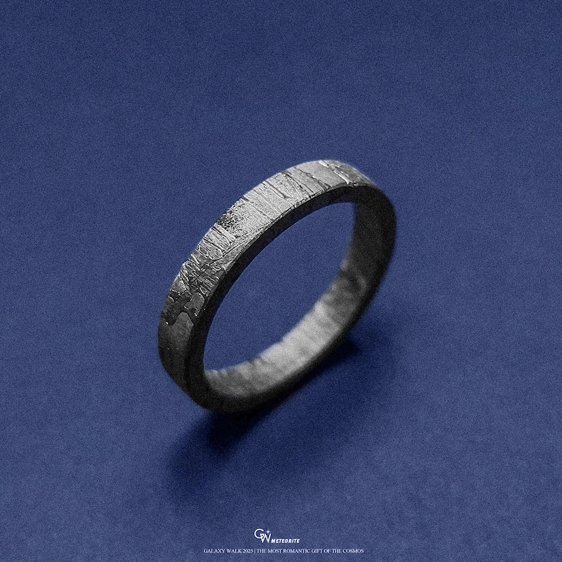 May include: A dark grey ring with a textured surface and a distinctive pattern, set against a deep blue backdrop. The ring's design has a unique, almost metallic appearance. The ring is a piece of jewellery.