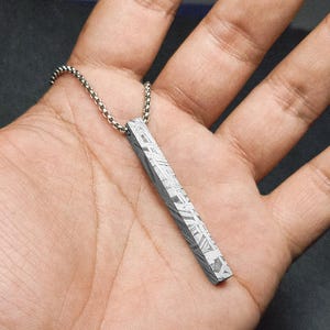 Peut inclure: Un collier avec un pendentif argenté en forme de barre rectangulaire. Le pendentif présente une surface à motifs distinctifs et est suspendu à une fine chaîne. Le design présente un motif géométrique. Le collier est présenté sur un fond neutre.