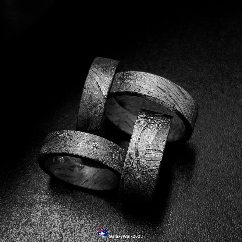 K&ouml;nnte beinhalten: Vier dunkelgraue Ringe mit einem strukturierten Muster werden vor einem schwarzen Hintergrund gezeigt. Die Ringe bestehen aus dunklem Metall und haben eine einzigartige, gemusterte Oberfl&auml;che. Die Ringe sind gestapelt und bilden eine visuell interessante Komposition.