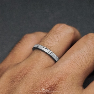 May include: A close-up of a ring with a distinctive, textured pattern. The ring is silver and grey, with a band that appears to be made of a stone-like material. The ring is on a finger, and the background is a dark grey.
