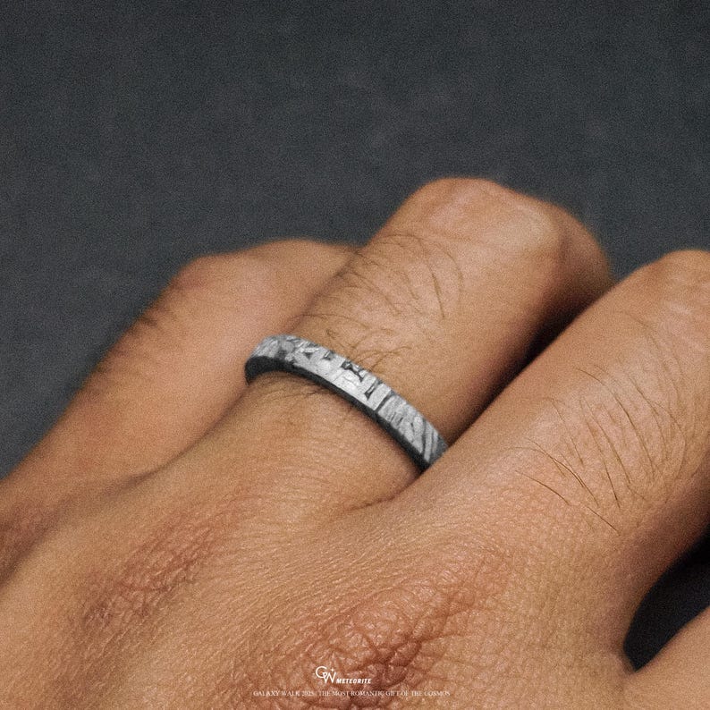 May include: A close-up of a ring with a distinctive, textured pattern. The ring is silver and grey, with a band that appears to be made of a stone-like material. The ring is on a finger, and the background is a dark grey.