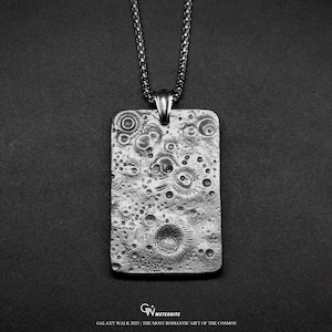May include: A silver-toned rectangular pendant with a textured surface, resembling a lunar landscape, hangs from a matching chain. The pendant features craters and other surface details. The text "GALAXY WALK 2025 | THE MOST ROMANTIC GIFT OF THE COSMOS" is at the bottom.