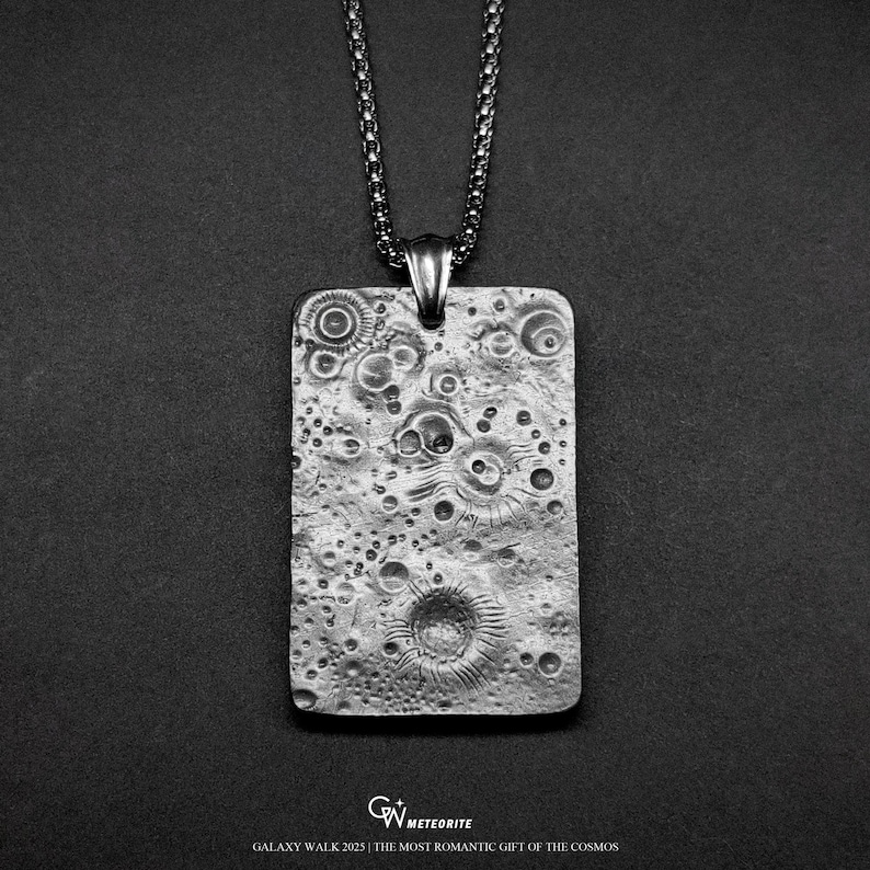 May include: A silver-toned rectangular pendant with a textured surface, resembling a lunar landscape, hangs from a matching chain. The pendant features craters and other surface details. The text "GALAXY WALK 2025 | THE MOST ROMANTIC GIFT OF THE COSMOS" is at the bottom.