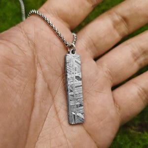 Op de afbeelding: Een zilveren ketting met een rechthoekige hanger gemaakt van meteoriet. De hanger heeft een uniek patroon van lijnen en swirls.