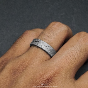 May include: A silver ring with a textured surface and geometric patterns. The ring is worn on a finger, showcasing its intricate design. The ring's surface has a unique, etched appearance, adding to its visual appeal. The ring is a piece of jewellery.