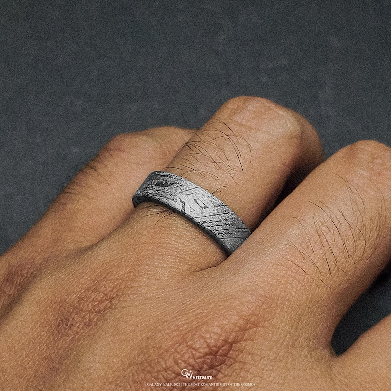 May include: A silver ring with a textured surface and geometric patterns. The ring is worn on a finger, showcasing its intricate design. The ring's surface has a unique, etched appearance, adding to its visual appeal. The ring is a piece of jewellery.
