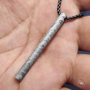 Puede incluir: Un collar con colgante que presenta un colgante cilíndrico de piedra gris con un diseño único. El colgante está suspendido de una cadena negra. El collar se muestra sobre un fondo neutro.