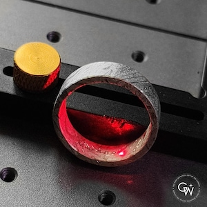 May include: Close-up of a silver ring with a textured pattern and a red interior. A small, gold-coloured cylindrical object is to the left of the ring. The ring is on a dark surface.