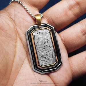 Puede incluir: Un colgante rectangular con un centro metálico estampado, enmarcado por bordes plateados y negros, y una fianza dorada. El colgante cuelga de una cadena plateada. El texto "GALAXY WALK 2023 THE MOST ROMANTIC GIFT OF THE COSMOS" está en la parte inferior.