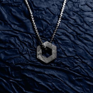 Puede incluir: Un collar de cadena de plata con un colgante hexagonal. El colgante tiene un diseño geométrico y es texturizado.
