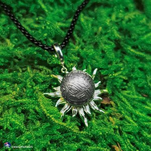 Könnte beinhalten: Silberne Anhänger-Halskette mit einem strukturierten Globus-Design, umgeben von Sonnenstrahlen. Der Anhänger hängt an einem schwarzen Band. Das Schmuckstück liegt auf grünem Moos.