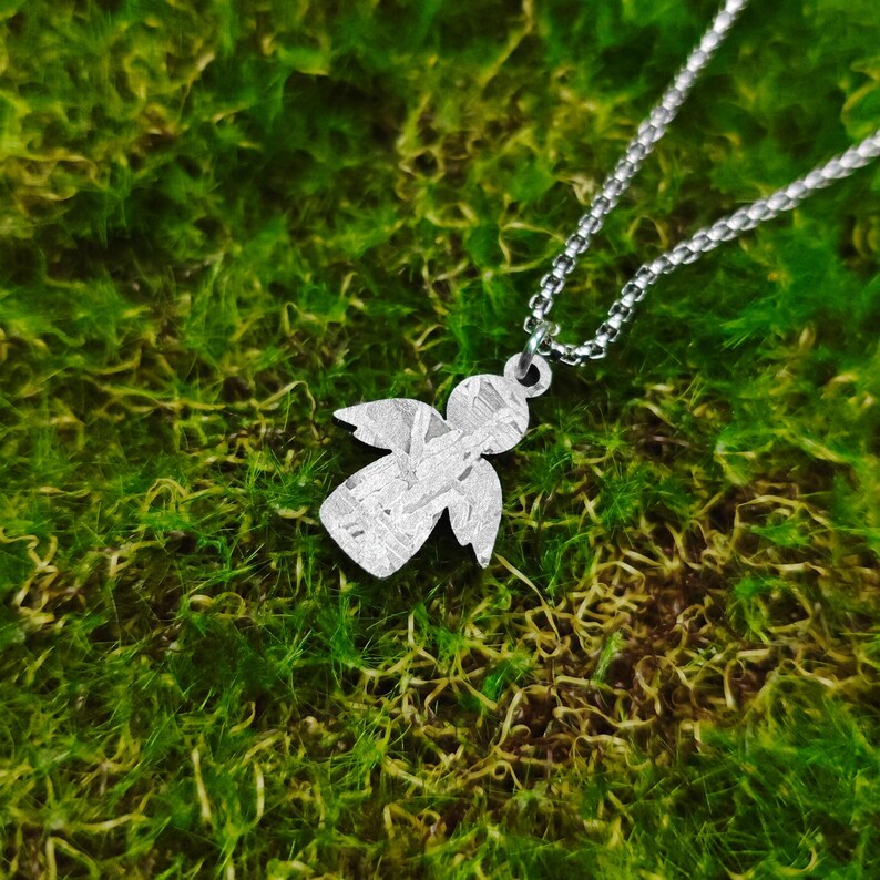 May include: A silver angel pendant necklace with a textured finish, hanging on a silver chain against a green moss background.