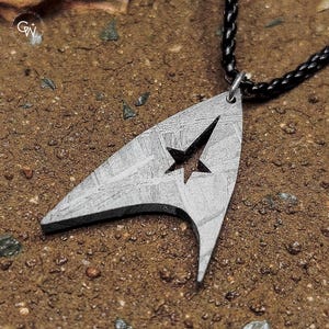 May include: A silver pendant shaped like a Starfleet emblem, featuring a star cut-out, suspended from a black, braided cord necklace. The background is a textured, earthy surface.