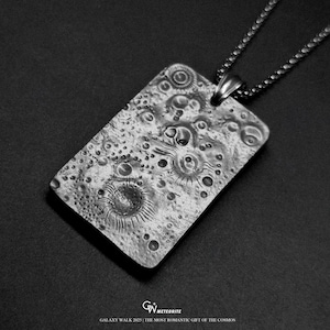 May include: A rectangular silver pendant with a textured surface, resembling a lunar landscape, hangs from a chain. The pendant features craters and circular patterns. The text "GALAXY WALK 2025 | THE MOST ROMANTIC GIFT OF THE COSMOS" is at the bottom.