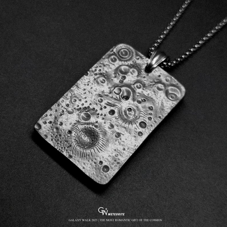 May include: A rectangular silver pendant with a textured surface, resembling a lunar landscape, hangs from a chain. The pendant features craters and circular patterns. The text "GALAXY WALK 2025 | THE MOST ROMANTIC GIFT OF THE COSMOS" is at the bottom.