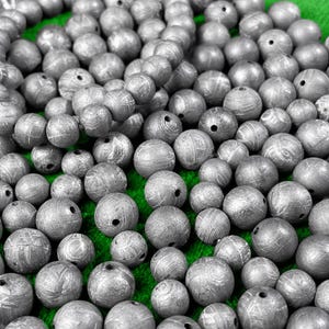Può includere: Primo piano di un mucchio di perline di metallo color argento. Le perline sono rotonde e hanno una superficie testurizzata. Sono disposte in uno schema casuale su una superficie verde.