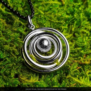 May include: A silver-toned pendant featuring a central textured sphere surrounded by concentric circular rings, suspended from a black chain. The text at the bottom reads: GALAXY WALK | THE MOST ROMANTIC GIFT OF THE COSMOS.