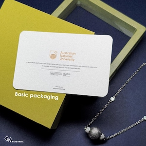 May include: A white rectangular card with text from the Australian National University rests on a yellow box. The card states it is a meteorite certificate. A silver necklace with a dark grey bead and clear stones is also visible.