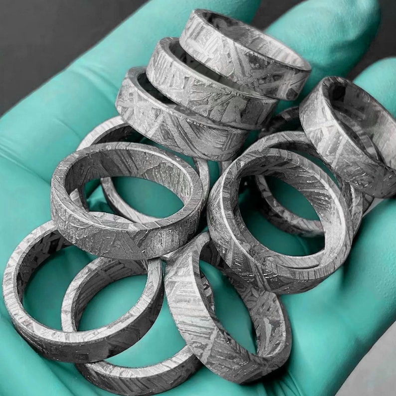 K&ouml;nnte beinhalten: Eine Ansammlung silberfarbener Ringe mit einem einzigartigen Muster. Die Ringe sind gestapelt, wodurch ihre komplizierten Details hervorgehoben werden. Die Ringe scheinen aus Metall zu sein und werden vor einem t&uuml;rkisfarbenen Hintergrund pr&auml;sentiert.