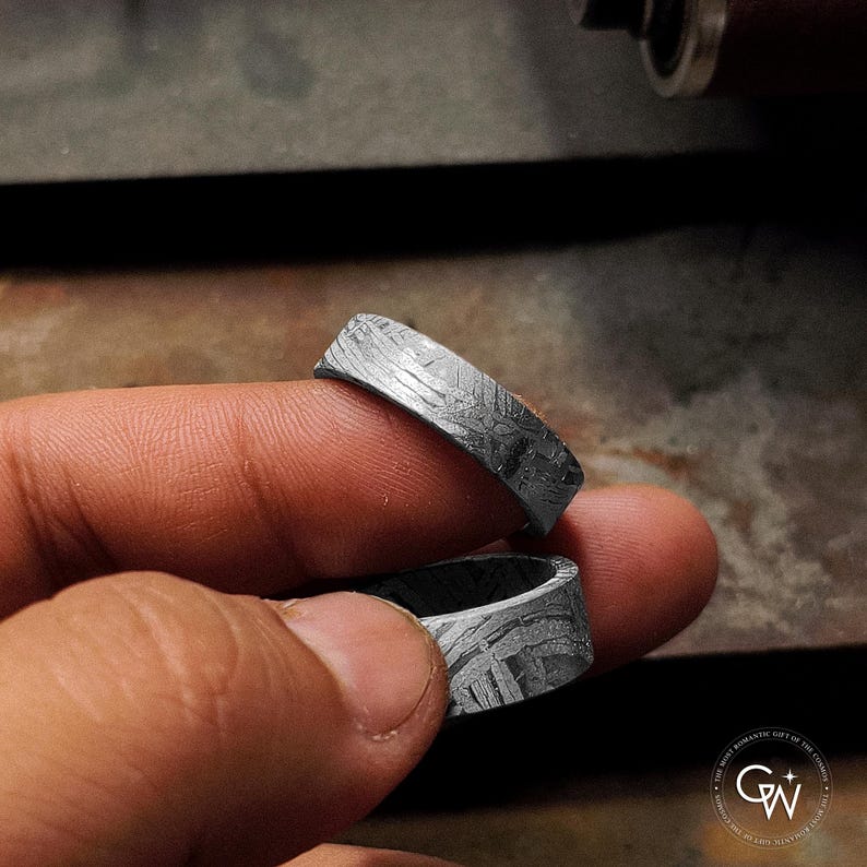K&ouml;nnte beinhalten: Zwei silberne Ringe mit strukturiertem Muster werden zwischen Fingern gehalten. Die Ringe haben ein dunkles, metallisches Aussehen und eine einzigartige, gemusterte Oberfl&auml;che. Die Ringe sind wahrscheinlich Teil einer Schmuckkollektion.