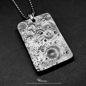 May include: A rectangular silver pendant with a textured surface, resembling a celestial body, hangs from a chain. The pendant features craters and circular designs. The text "GALAXY WALK 2025 / THE MOST ROMANTIC GIFT OF THE COSMOS" is at the bottom.