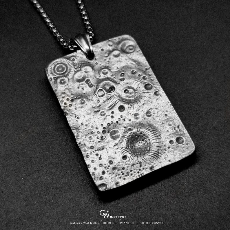 May include: A rectangular silver pendant with a textured surface, resembling a celestial body, hangs from a chain. The pendant features craters and circular designs. The text "GALAXY WALK 2025 / THE MOST ROMANTIC GIFT OF THE COSMOS" is at the bottom.