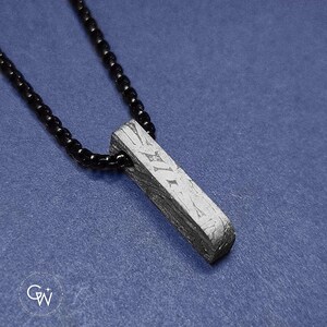 Puede incluir: Un collar con colgante rectangular y cadena de cuentas negras. El colgante es de metal gris texturizado con marcas grabadas. El collar se muestra sobre un fondo azul.