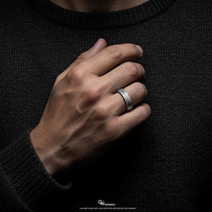 May include: A silver ring with an etched pattern. The ring is worn on a finger, displaying the detailed design. The ring's surface reflects light, highlighting the texture and craftsmanship. The background is a dark, textured fabric.