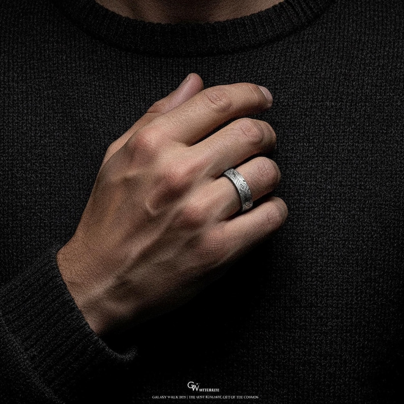 May include: A silver ring with an etched pattern. The ring is worn on a finger, displaying the detailed design. The ring's surface reflects light, highlighting the texture and craftsmanship. The background is a dark, textured fabric.