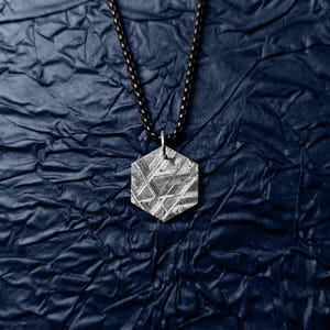 Puede incluir: Un collar con un colgante hexagonal de plata en una cadena negra. El colgante tiene una superficie texturizada con un patrón de líneas.