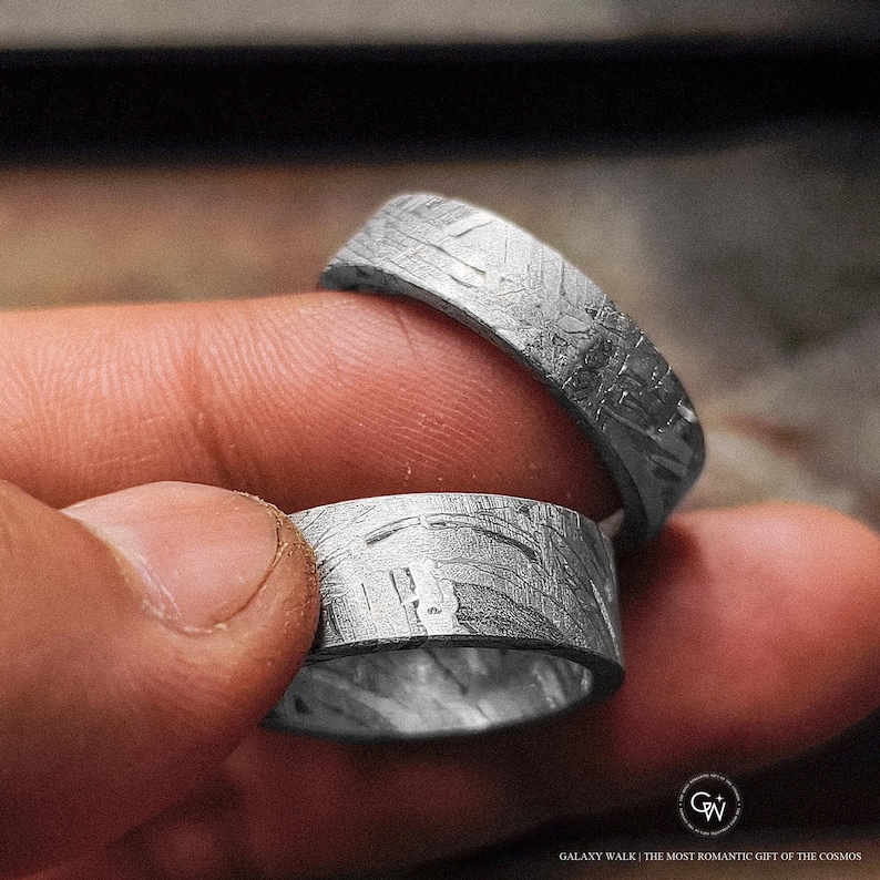 May include: Two silver rings with a textured, patterned surface. The rings are held between fingers, showcasing their unique design. The rings have a band-like shape and appear to be made of metal.