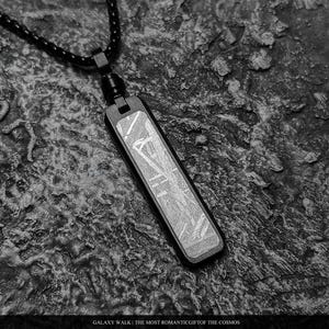 Puede incluir: Un collar con colgante rectangular que presenta un marco de metal negro y una incrustación metálica texturizada con un patrón entrecruzado. El colgante cuelga de una cadena negra. El texto en la parte inferior dice: GALAXY WALK | THE MOST ROMANTIC GIFT OF THE COSMOS.