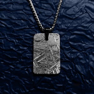 Puede incluir: Un colgante rectangular con una superficie metálica texturizada cuelga de una cadena plateada. El colgante tiene un diseño oscuro y estampado. El collar se muestra sobre un fondo azul oscuro texturizado.