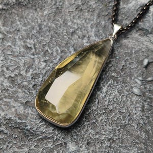 Könnte beinhalten: Ein tropfenförmiger Anhänger mit einem facettierten, durchscheinenden gelb-grünen Edelstein in einer silberfarbenen Metallfassung. Der Anhänger hängt an einer dunklen, strukturierten Metallkette und weist eine polierte Oberfläche auf.