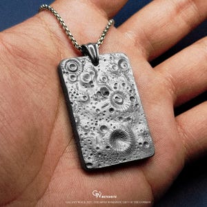 May include: A rectangular pendant with a textured surface resembling a lunar landscape, suspended from a silver chain. The pendant features craters and other surface details. The text "GALAXY WALK 2025 / THE MOST ROMANTIC GIFT OF THE COSMOS" is visible.