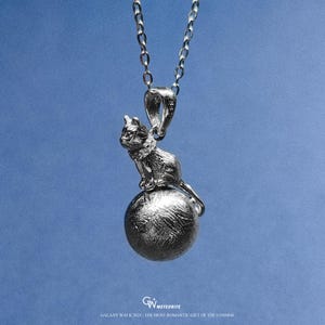 Puede incluir: Un collar con colgante plateado que representa un gato sentado sobre una esfera texturizada. El gato está detallado con pelo y rasgos faciales visibles. La cadena es de estilo eslabón delicado. El texto en la parte inferior dice "GALAXY WALK 2025 | THE MOST ROMANTIC GIFT OF THE COSMOS."