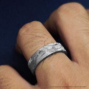 K&ouml;nnte beinhalten: Ein silberner Ring mit einem einzigartigen, strukturierten Muster wird an einem Finger getragen. Die Oberfl&auml;che des Rings zeigt ein detailliertes, unregelm&auml;&szlig;iges Design. Der Text "GALAXY WALK 2025 | THE MOST ROMANTIC GIFT OF THE COSMOS" ist unten sichtbar.