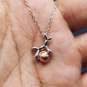 Peut inclure: Collier argenté avec un pendentif représentant un animal ballon tenant une sphère texturée de couleur cuivrée. La chaîne est fine et le pendentif est détaillé. Le collier est présenté sur un fond de couleur chair.