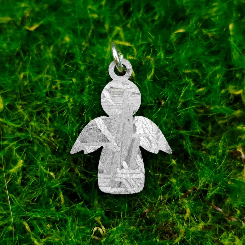 May include: A silver angel charm with a textured surface. The charm is hanging from a green mossy background.